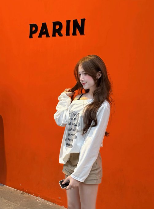 A young woman with long brown hair wearing a white blouse and tan shorts posing against a bright orange wall with the text 'PARIN' in black letters.