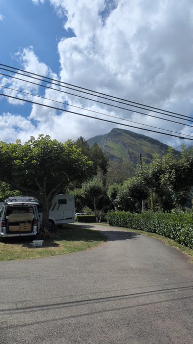 A camper van and caravan parked under leafy trees at a mountain campsite, with the green peaks of the Spanish Pyrenees rising beneath a dramatic cloudy sky—perfectly matching the post's "touching grass" getaway vibe before Sunday's BTC update.