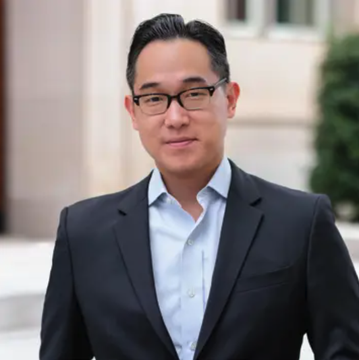 A professional headshot of an Asian man wearing glasses, a light blue button-down shirt, and a dark navy blazer, standing outdoors against an architectural backdrop, accompanying an article about crypto policy featuring commentary from the Crypto Council for Innovation CEO Ji Kim.
