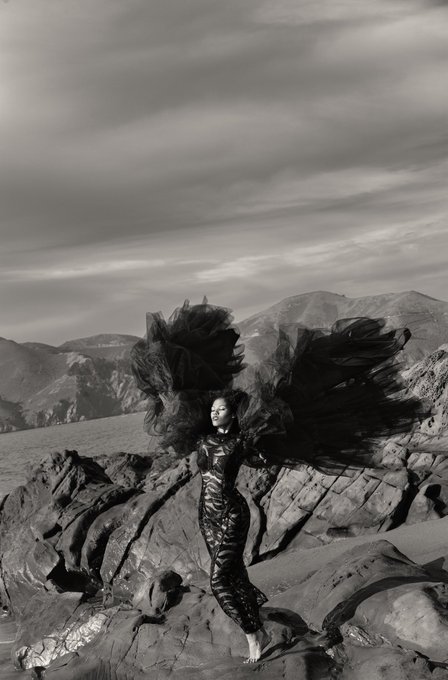 A black-and-white photograph shows a woman in a flowing dark dress with dramatic, wing-like fabric billowing around her, standing barefoot on rocky terrain against a mountainous landscape.