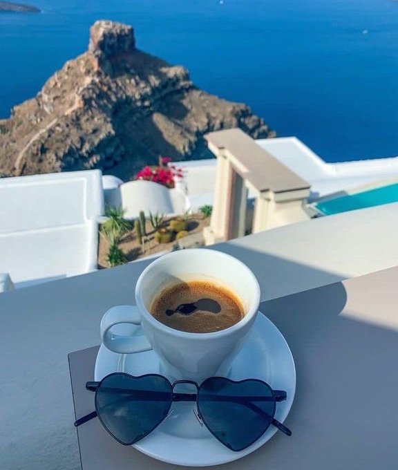 A scenic view of an espresso coffee on a saucer with heart-shaped sunglasses placed on it, overlooking a white cliffside terrace with the sea and a rocky promontory in the background, reminiscent of Santorini.