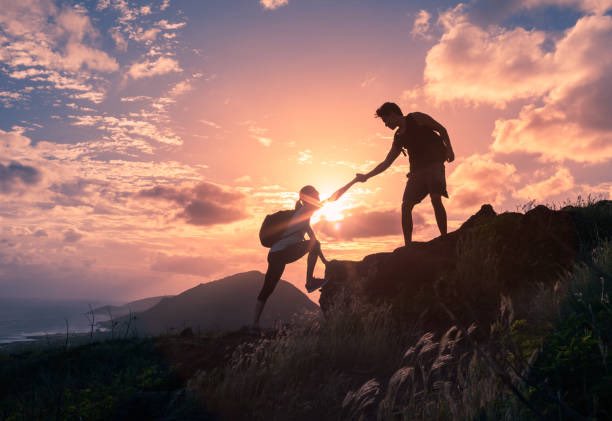 A silhouette photograph of two hikers, one helping the other up a rocky hill against a vibrant orange and pink sunset sky.