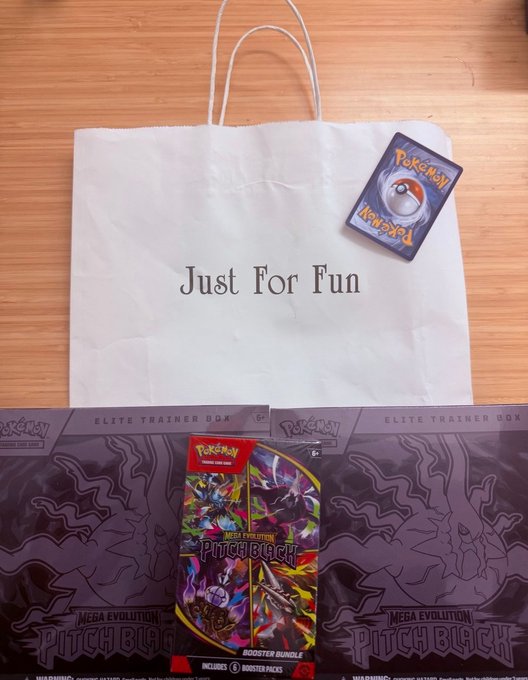 A flat-lay photograph showing a white shopping bag labeled 'Just For Fun' alongside two Pokémon TCG 'Mega Evolution Pitch Black' Elite Trainer Boxes and a booster bundle on a wooden surface.