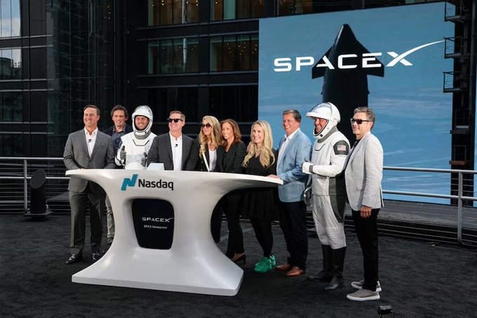 A group photo of SpaceX employees, including those in space suits, standing at a Nasdaq podium with a SpaceX banner behind them, celebrating a SpaceX milestone.