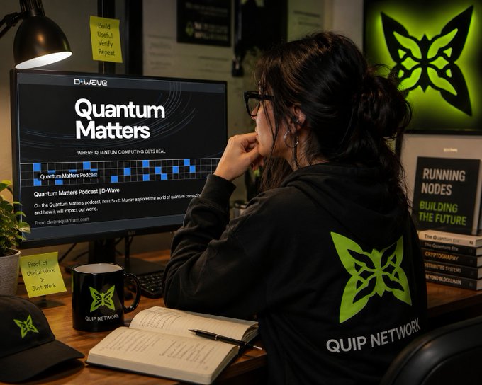 A person wearing a 'QUIP NETWORK' hoodie sits at a desk looking at a monitor displaying 'D-Wave Quantum Matters', with branded merchandise scattered around the workspace.