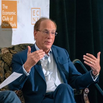 Man in a blue blazer gesturing while speaking at an event for The Economic Club of New York (ECNY), with the club's logo visible on a banner in the background.