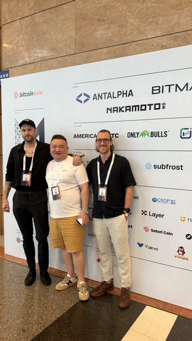 Three men pose together in front of a sponsor backdrop at a crypto event, with visible logos including 'bitcoin asia', 'ANTALPHA', 'NAKAMOTO', 'subfrost', 'Layer', and 'Satomi Coin'.