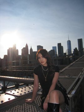 A young woman with a yellow headband and black outfit sits on a metal grate rooftop structure with the New York City skyline, including the Freedom Tower, visible in the bright background.