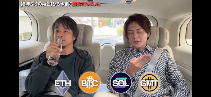 The image shows two men sitting inside a car, with one of them drinking water from a bottle while the other man looks at him. The background features a gray wall that has various logos displayed on it, including "Ethereum", "Btc", "SOL", and "GMT".