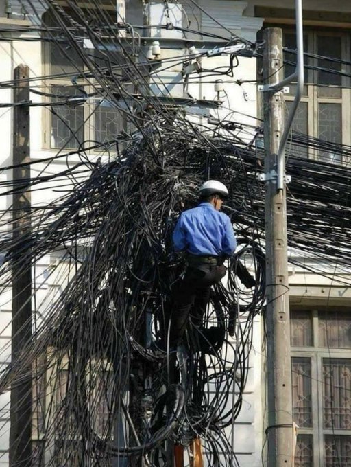 A worker in a blue shirt and white helmet climbs a utility pole heavily tangled in a massive, chaotic bundle of black wires in front of a building.
