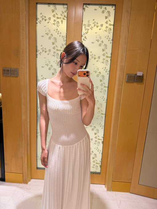 A woman takes a mirror selfie wearing a sheer white dress with a square neckline, holding a smartphone in a room with floral-patterned glass doors.