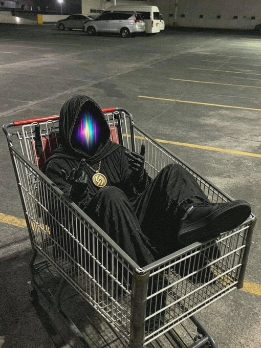 An artistic image of a hooded figure wearing black robes and a gold pendant, with a glowing colorful light beam emanating from the hood instead of a face, sitting in a shopping cart in a parking lot at night.