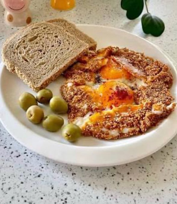A breakfast plate contains fried eggs with runny yolks, two slices of brown bread, and a side of green olives, with a glass of orange juice and a small figurine in the background.