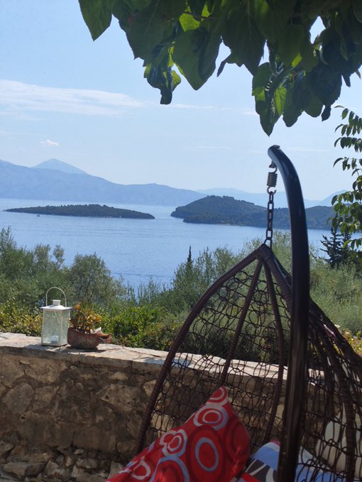 A scenic view from a stone terrace featuring a hanging wicker chair with a red patterned cushion, overlooking a blue sea with small islands and mountains in the distance.
