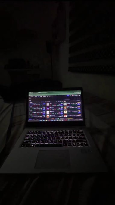 A dark bedroom scene with a glowing laptop on a bed, displaying what appears to be a live streaming interface with various icons and chat windows.