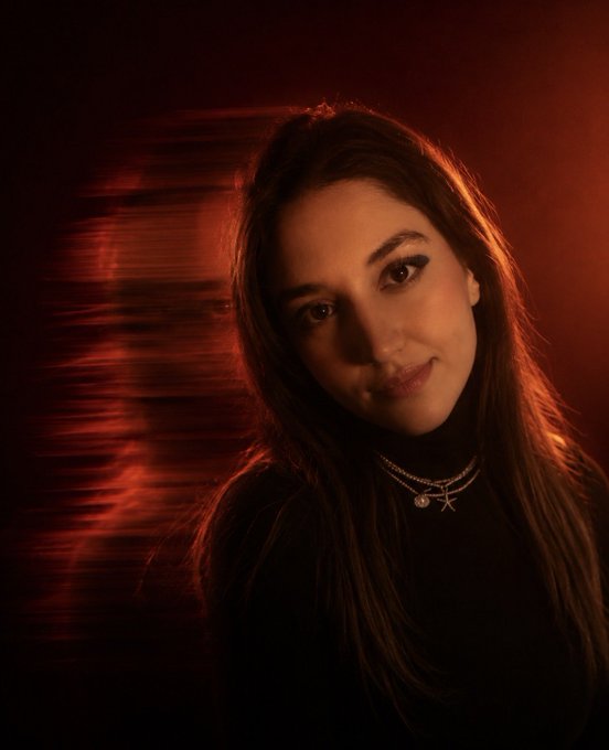A portrait of a young woman with long dark hair, wearing layered necklaces, photographed in moody red lighting with a blurred reflection effect.