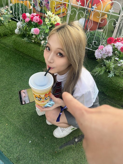 A young woman with blonde hair drinking from a large plastic cup with a straw, sitting on artificial grass next to colorful flowers while holding a smartphone.