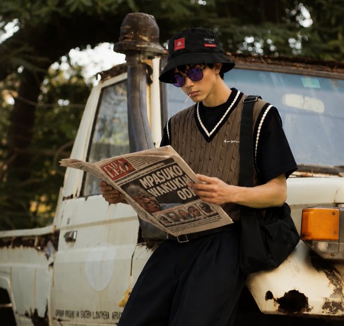 A young man in a bucket hat, purple sunglasses, and a black knit vest reads a newspaper featuring the headline 'MPASUKO WAIKWA OD' while leaning against a white vehicle.