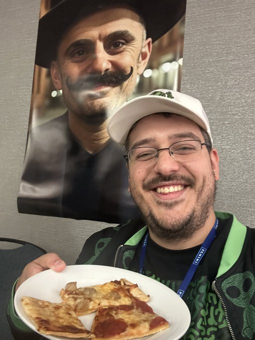 A smiling man wearing glasses and a white cap holds a plate of pizza slices, sitting in front of a large portrait of a man with a curled mustache.