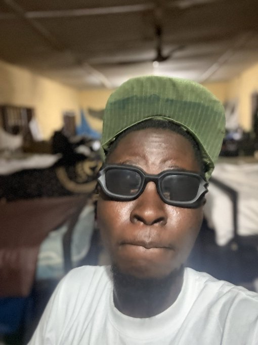 A close-up selfie of a man wearing dark rectangular glasses and a green backwards cap in an indoor room.