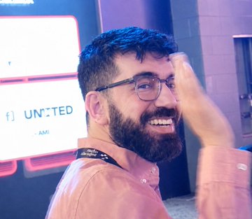 A photograph of a smiling bearded man wearing glasses and a pink/salmon shirt, posing in front of a sign that reads 'UNITED' and 'MIAMI.'