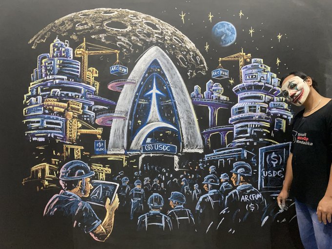 An artist wearing a Guy Fawkes mask and a "Saudi Standup" t-shirt stands next to a large black painting depicting a futuristic lunar city with "USDC" and "ARC FUN" signs, construction cranes, and an Earth visible in the starry sky.