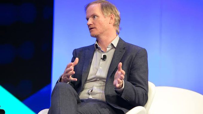 A man in a dark blazer and light blue shirt sits on a white chair against a blue backdrop, gesturing with both hands as he speaks during what appears to be a panel discussion, likely addressing BitGo's $42.5M acquisition of NYDIG's trading arm.