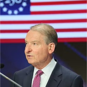 An older man in a dark suit with a purple tie speaks at a podium with an American flag and blue backdrop behind him, likely depicting a U.S. financial regulator (SEC or CFTC) addressing the topic of expedited crypto rulemaking in relation to the Clarity Act and $XRP.