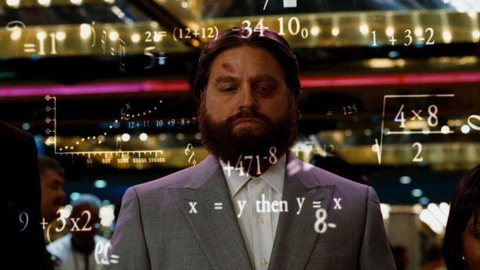 A bearded man in a suit standing in a brightly lit environment with mathematical equations and formulas floating around him.