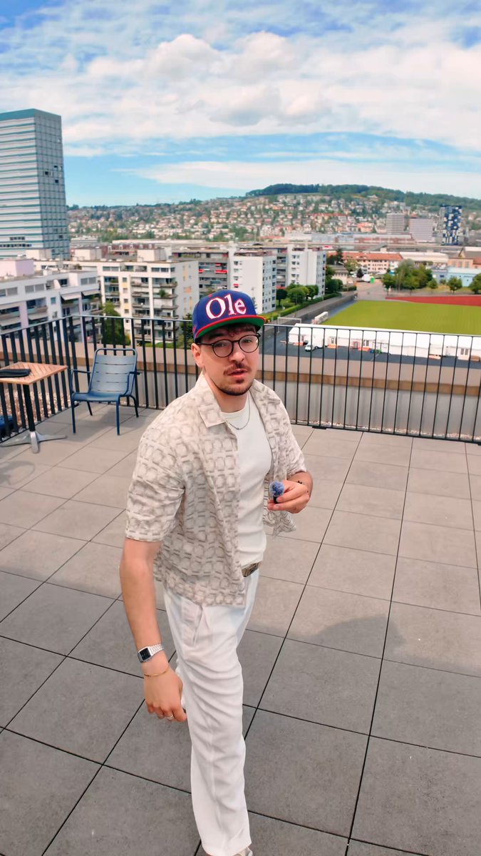 The image shows a man wearing glasses, white pants, and a hat standing on a rooftop or patio area with a city skyline in the background. The man has his arm outstretched towards the camera, as if posing for a photo. There is also an object near him that appears to be a cell phone. In the post, it seems like someone has posted a picture of the man and the surrounding environment on social media or another platform with a caption about connecting with an agent via a messaging app. The image focuses on the man's outfit and his connection to the agent through the use of technology.