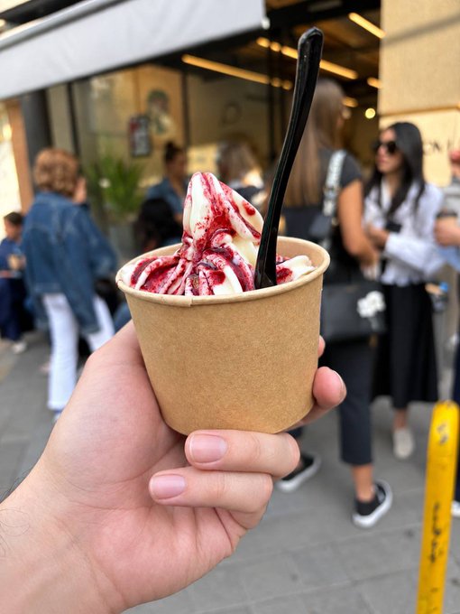 A hand holds a brown paper cup filled with swirled white soft-serve ice cream drizzled with red berry sauce and a black spoon, with blurred people on a city street in the background.