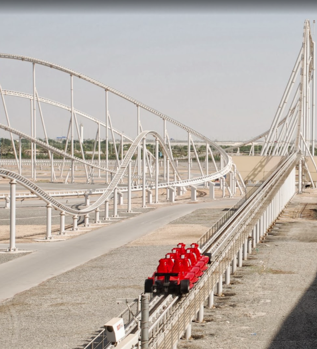 A red roller coaster train on a track approaching a massive white steel structure featuring multiple loops and curves in a desert-like setting.