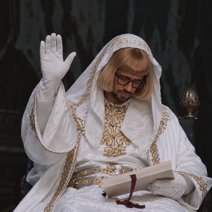 A man dressed in elaborate white and gold robes with a veil and white gloves reads from a book, raising one hand.