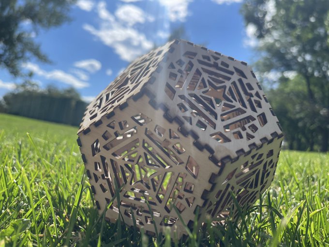 A complex, laser-cut geometric wooden sculpture resting on green grass under a bright blue sky with scattered clouds.