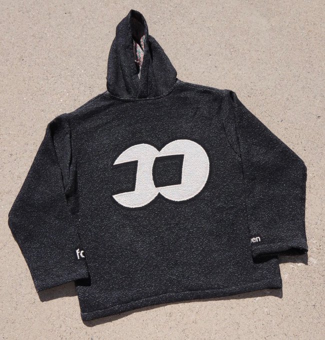 A black hoodie with a speckled texture is laid flat on the ground, featuring a large white geometric logo on the chest and small text on the sleeves.