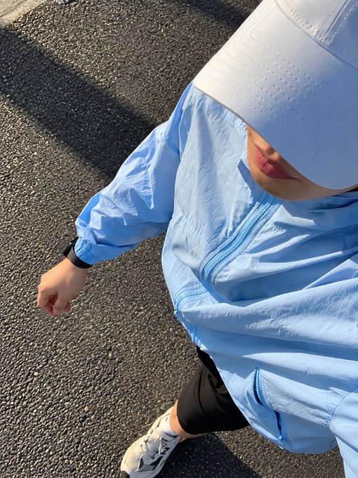 A selfie-style overhead shot of a person wearing a white cap pulled down over their eyes and a light blue windbreaker jacket, standing on asphalt pavement with their fist extended toward the camera.