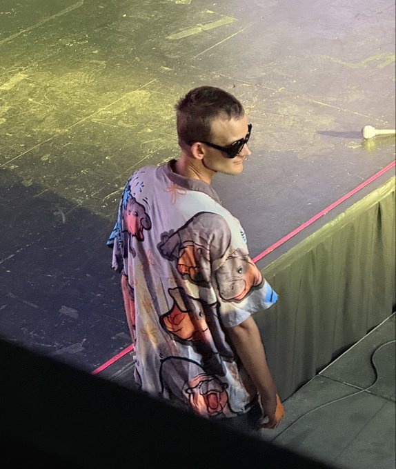 A young man with short hair and dark sunglasses stands backstage in a brightly lit venue, wearing an oversized cartoon-printed shirt. The image appears to be a casual backstage shot, likely from a livestream or event setting, but shows no visible text, logos, or branding related to "ResupplyFi."