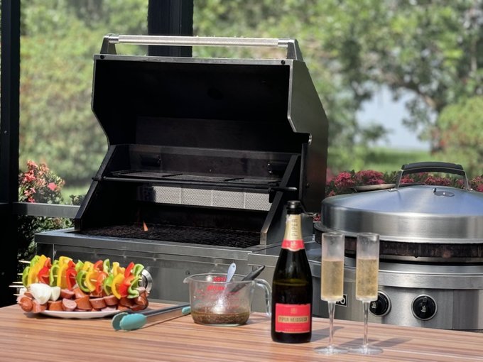 An outdoor patio setup featuring an open stainless steel grill alongside a plate of skewered meat and vegetables, champagne glasses, a bottle, and sauce, with a lake and trees in the background.