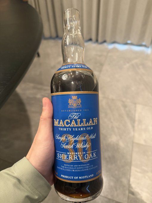 A hand holds up a bottle of The Macallan Thirty Years Old Single Highland Malt Scotch Whisky, showcasing its distinctive blue label.