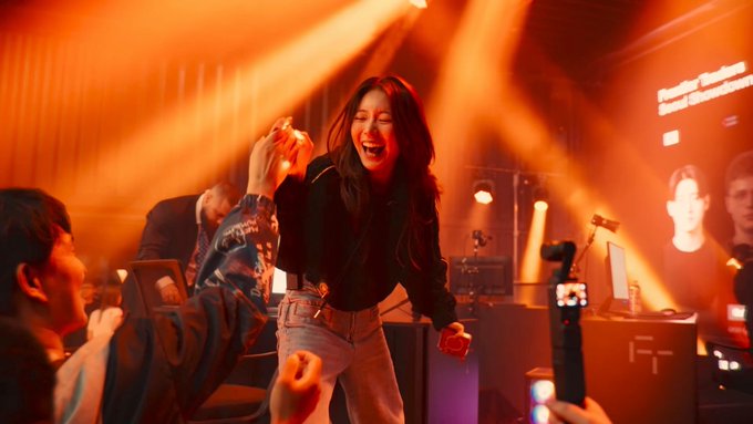 A woman laughs joyfully while dancing in a venue illuminated by dramatic orange stage lights, with audience members filming her on their phones.