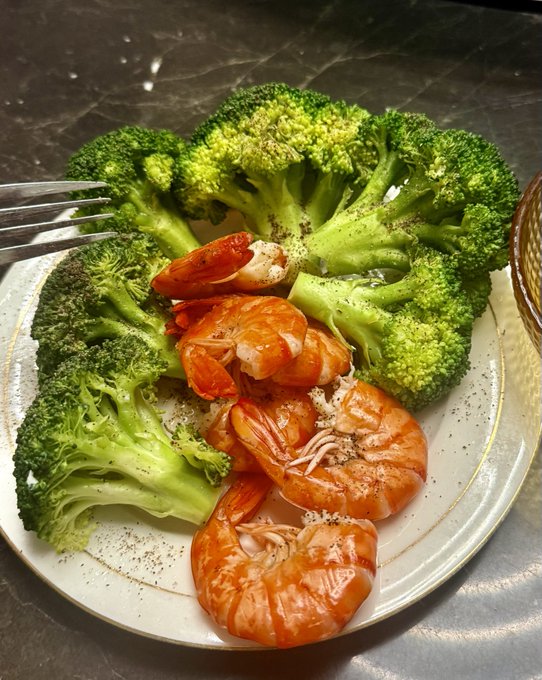 A close-up photo of a meal on a white plate features a generous serving of bright green steamed broccoli topped with several orange cooked shrimp and a metal fork resting on the edge.
