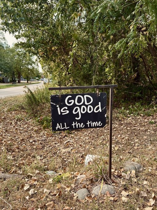 A handmade black sign stands in a yard covered with fallen leaves, reading 'GOD is good All the time' in white lettering, with a street visible in the background.