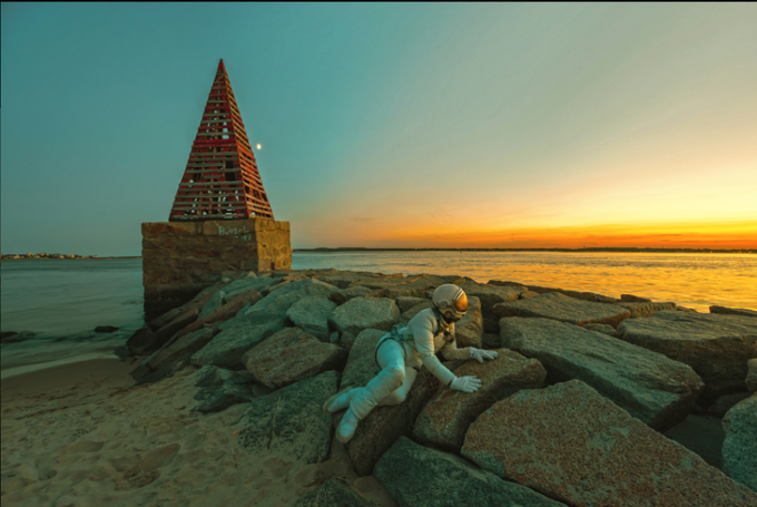 An astronaut in a white suit crawls across large rocks on a sandy beach near a red pyramid-shaped sculpture, with a golden sunset over the water in the background.