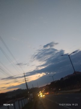 A dusk photograph taken with a Redmi 15 phone (timestamp 11/07/2026 19:23) showing a road with a car and streetlights under a cloudy sky.