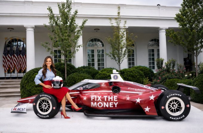 A woman in a blue blouse and red skirt sits on a red race car displaying 'FIX THE MONEY' in front of a white-columned building with an American flag.