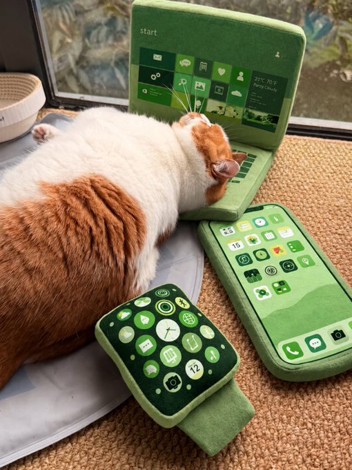 An orange and white cat lies peacefully on miniature green laptop, smartphone, and smartwatch toys designed to look like Apple's devices with green-themed app icons.
