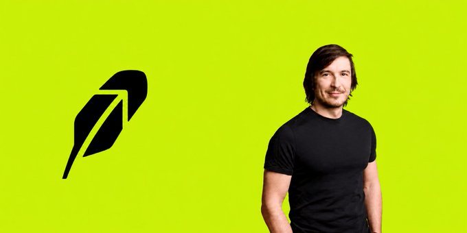 A man with brown hair wearing a black t-shirt standing next to a black bird-like logo on a bright yellow-green background.