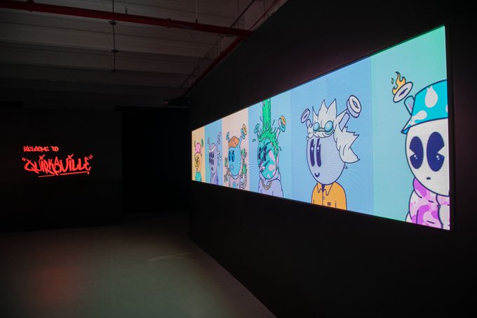 An art gallery installation featuring a row of large digital screens displaying colorful cartoon character NFTs, with a red neon sign reading 'WELCOME TO QUINTVILLE' on the left wall.
