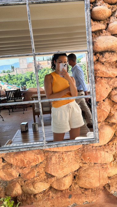 A person takes a mirror selfie wearing an orange tank top and white shorts, reflecting a scenic view with a stone fortress and lush greenery in the background.