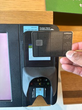 The image shows a person holding a credit card reader with a black screen, which appears to be an Amex Gold card reader. The card reader has a small screen that displays information about the card, such as its expiration date or balance. The card is being used for payment at a store, and it is being held up by someone's hand.